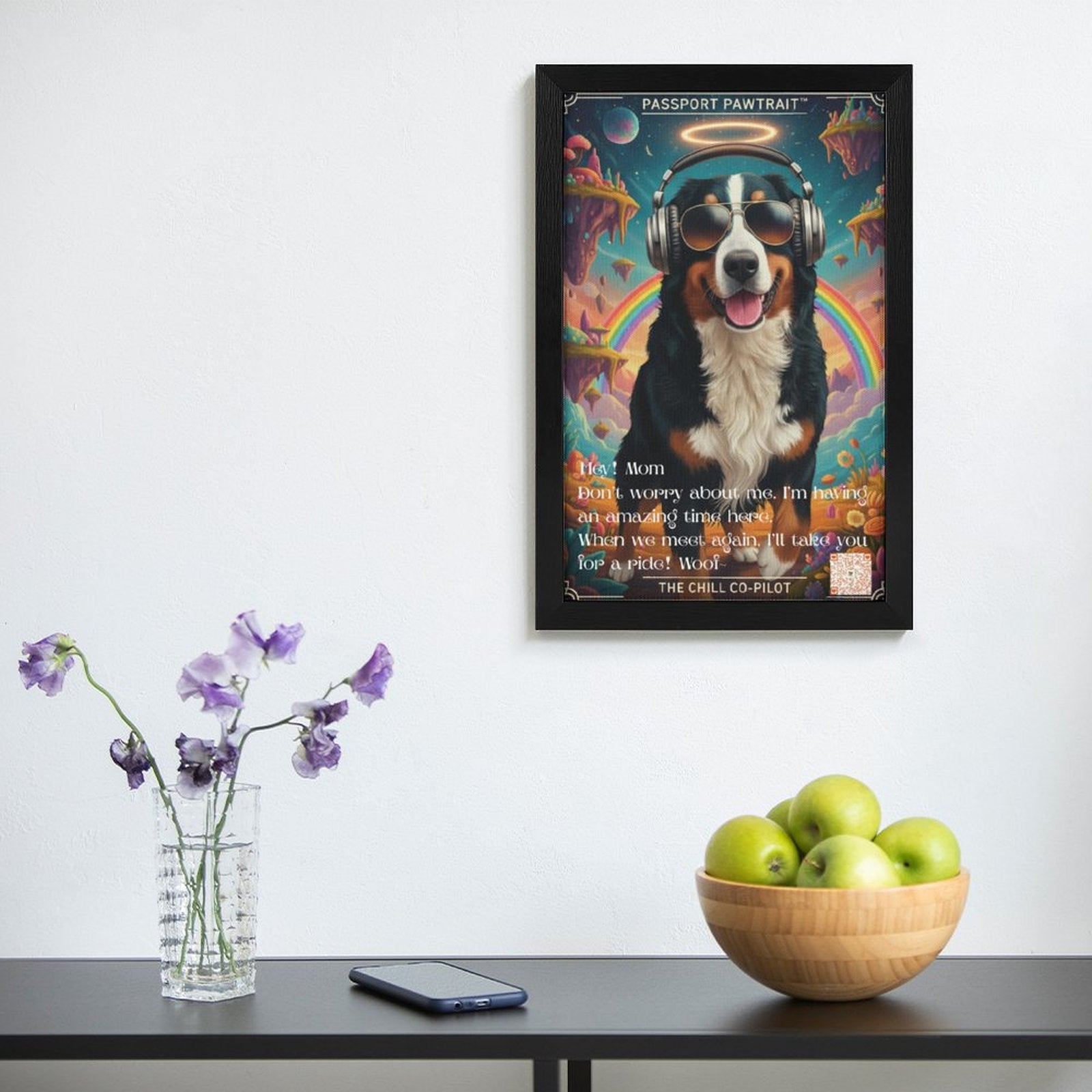 Framed artwork of a dog with headphones on a wall above a table with a vase, phone, and bowl of apples.